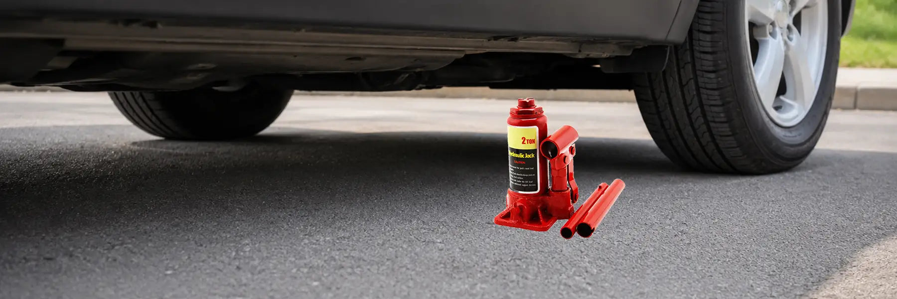 A red hydraulic bottle jack and extension handles resting on the pavement underneath the frame rail of a black passenger car.