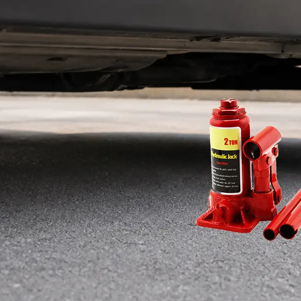 A red hydraulic bottle jack and extension handles resting on the pavement underneath the frame rail of a black passenger car.