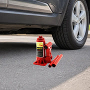 A red hydraulic bottle jack and extension handles resting on the pavement underneath the frame rail of a black passenger car.